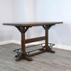 Wooden table with legs on a gray floor against a white wall. Homestead Pub Table available at Rustic Ranch Furniture and Decor.