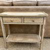 Wooden side table with two drawers and an open shelf in front of a brown sofa. Beach Pebble Side Table available at Rustic Ranch Furniture and Decor.