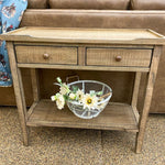 Wooden side table with two drawers and a wire basket with flowers on a carpeted floor. Beach Pebble Side Table available at Rustic Ranch Furniture and Decor.