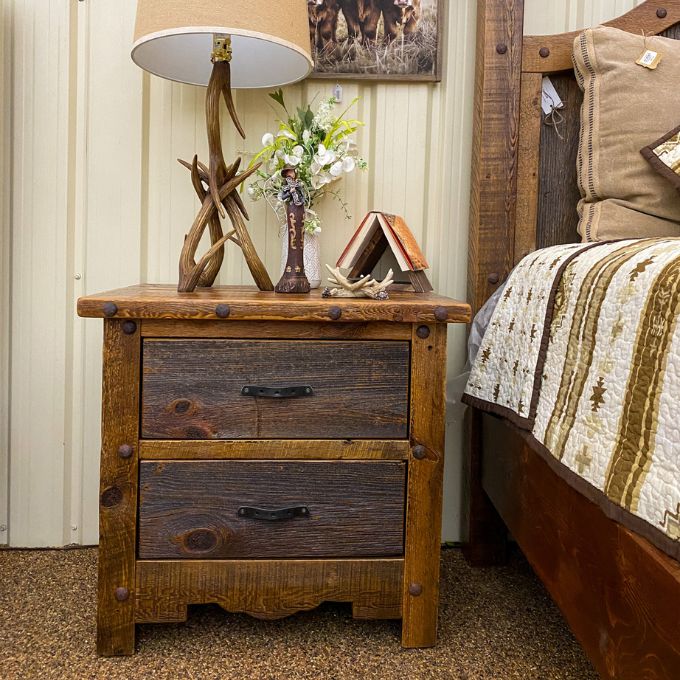 Wooden nightstand with lamp and decor next to a bed with patterned bedding. Black Hills Two Drawer Nightstand available at Rustic Ranch Furniture and Decor.