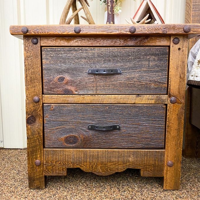 Wooden nightstand with two drawers and metal handles on a carpeted floor. Black Hills Two Drawer Nightstand available at Rustic Ranch Furniture and Decor.