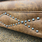 Close-up of a brown leather chair with metal studs on a textured background.  Emerson Chair and a Half available at Rustic Ranch Furniture and Decor.