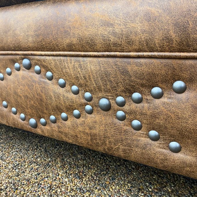Close-up of a brown leather chair with metal studs on a textured background.  Emerson Chair and a Half available at Rustic Ranch Furniture and Decor.