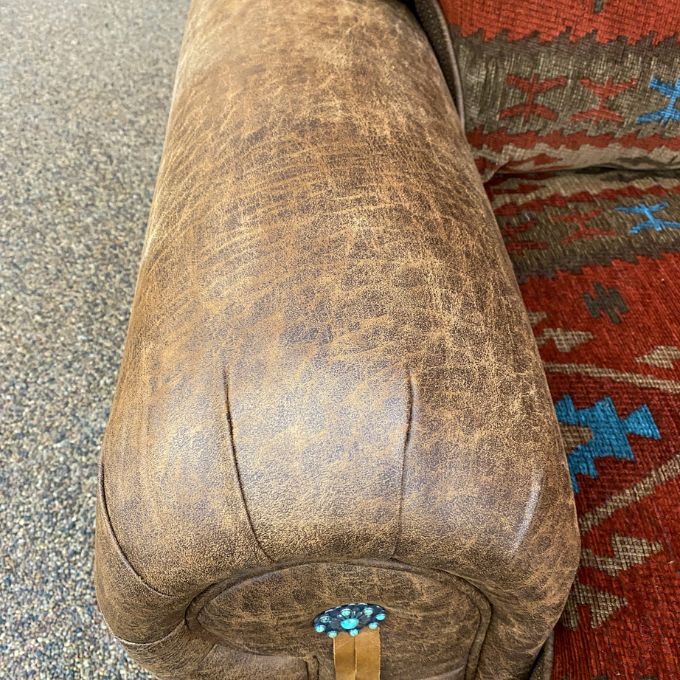 Brown leather ottoman with a decorative strap on a textured surface.  Emerson Chair and a Half available at Rustic Ranch Furniture and Decor.