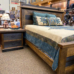 Wooden bed with bedding and pillows in a store setting. Fireside Nightstand available at Rustic Ranch Furniture and Decor.
