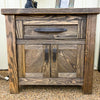 Wooden nightstand with a single drawer and two doors on a textured surface. Dutton Nightstand available at Rustic Ranch Furniture and Decor.