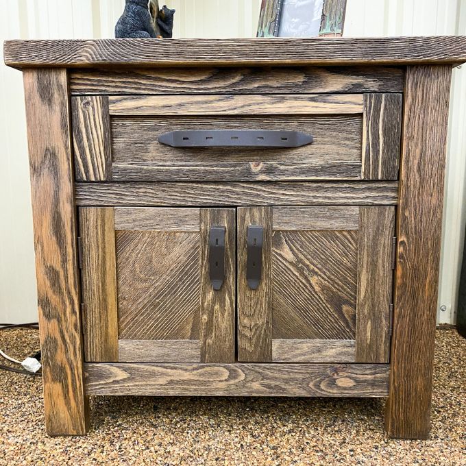 Wooden nightstand with a single drawer and two doors on a textured surface. Dutton Nightstand available at Rustic Ranch Furniture and Decor.
