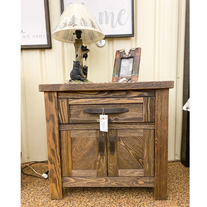 Wooden nightstand with a lamp and picture frame on top, in a room setting. Dutton Nightstand available at Rustic Ranch Furniture and Decor.
