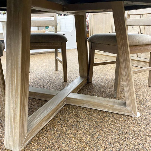 Wooden dining table base with chairs on a textured floor. Rowan Round Dining Table available at Rustic Ranch Furniture and Decor.