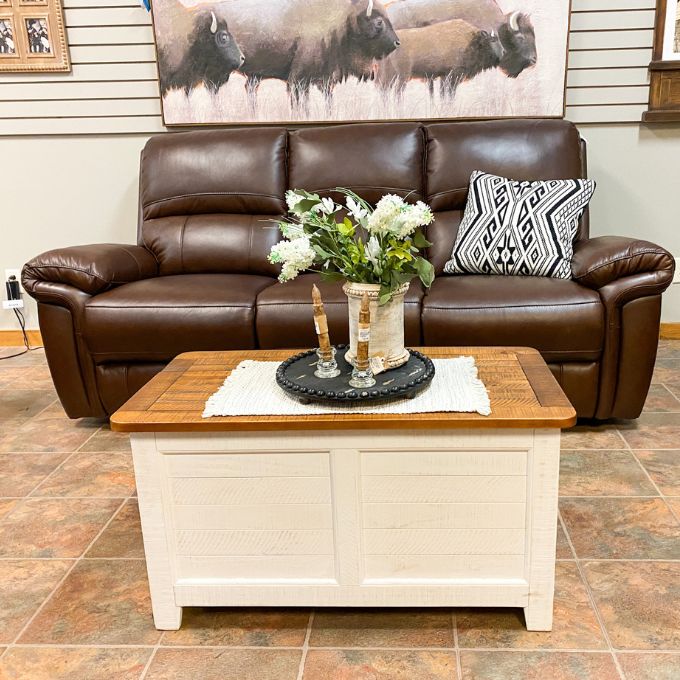 Brown leather sofa with a wooden coffee table in a room with a decorative wall piece. White Dashbury Storage Trunk available at Rustic Ranch Furniture and Decor.