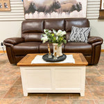 Brown leather sofa with a wooden coffee table in a room with a decorative wall piece. White Dashbury Storage Trunk available at Rustic Ranch Furniture and Decor.