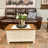 Brown leather sofa with a wooden coffee table in a room with a painting of bison on the wall. White Dashbury Storage Trunk available at Rustic Ranch Furniture and Decor.