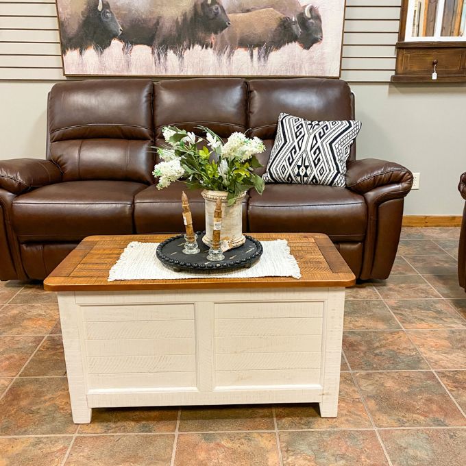Brown leather sofa with a wooden coffee table in a room with a painting of bison on the wall. White Dashbury Storage Trunk available at Rustic Ranch Furniture and Decor.