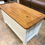 Wooden storage chest with a white base on a tiled floor. White Dashbury Storage Trunk available at Rustic Ranch Furniture and Decor.